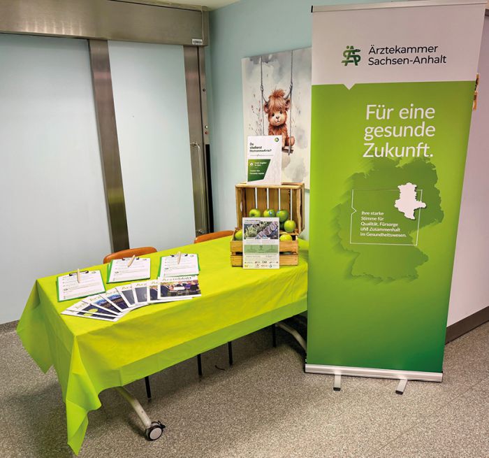 Sitzung der Ärztekammer mit zahlreichen Teilnehmern an Tischen, Vortragendem am Podium und Präsentation im Konferenzraum.