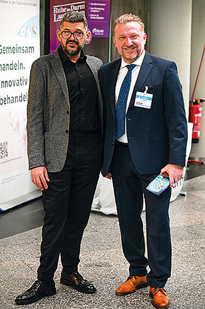 Zwei Männer im Anzug und Sakko stehen nebeneinander auf einem Kongress und lächeln in die Kamera.