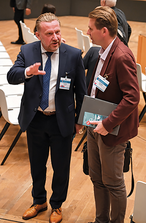 Zwei Männer im Gespräch auf einem Kongress; beide tragen Namensschilder, einer hält Unterlagen, der andere gestikuliert.